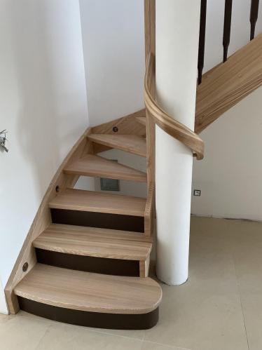 Spiralne schody z jasnego drewna z ciemnobrązowymi podstopnicami, wsparte na białym filarze, w jasnym wnętrzu. Widok z dołu na pierwsze stopnie.