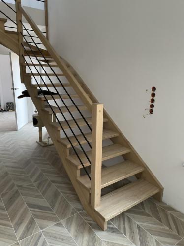 Jasne schody z drewna z czarną, minimalistyczną balustradą, ustawione na wzorzystej podłodze. Widoczne gniazdka elektryczne na ścianie.