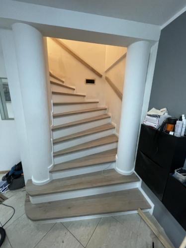 Spiralne schody w jasnym wnętrzu, z drewnianymi stopniami i białymi kolumnami. Widoczne detale wykończenia i oświetlenie.