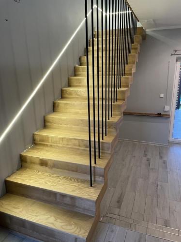Drewniane schody z czarną, minimalistyczną balustradą i subtelnym oświetleniem LED wzdłuż ściany. Jasne drewno stopni harmonizuje z szarą ścianą i podłogą.