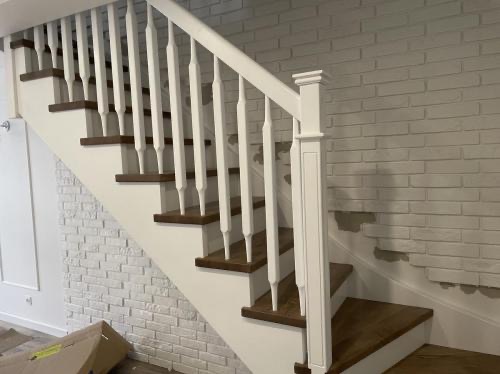 Klasyczne schody z drewnianymi stopniami i białą balustradą na tle ściany z białej cegły. Widok z dołu, skupiający się na detalach wykończenia.