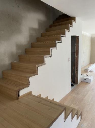 Montaż jasnych, drewnianych schodów w surowym wnętrzu. Widoczne niedokończone ściany i podłogi, podkreślają etap prac wykończeniowych.