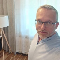 Tobiasz Szlęk - Agent nieruchomości - Portret mężczyzny w okularach na tle jasnego okna i zasłon, z lampą podłogową widoczną po lewej stronie. Drewniana podłoga dodaje ciepła wnętrzu.