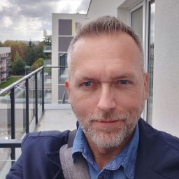 Tobiasz Szlęk - Agent nieruchomości - Mężczyzna na balkonie nowoczesnego apartamentowca, ubrany w denimową koszulę i marynarkę. W tle widoczne budynki mieszkalne i pochmurne niebo. Ujęcie z bliska.