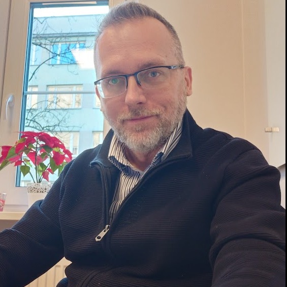 Portret mężczyzny w okularach, w tle okno z widokiem na budynek i poinsecja. Zdjęcie w neutralnym oświetleniu, z bliska, ukazujące twarz i górną część tułowia.