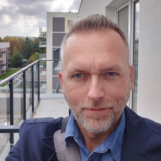 Mężczyzna na balkonie nowoczesnego apartamentowca, ubrany w denimową koszulę i marynarkę. W tle widoczne budynki mieszkalne i pochmurne niebo. Ujęcie z bliska.