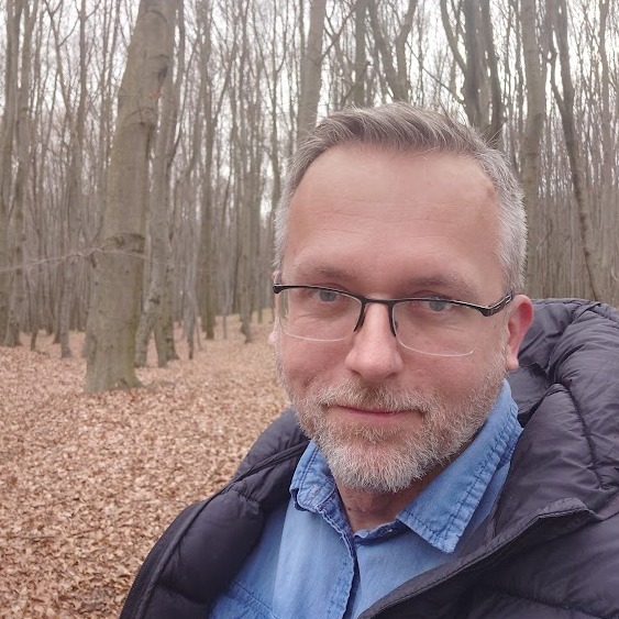 Portret mężczyzny w okularach na tle jesiennego lasu. Ubrany w dżinsową koszulę i kurtkę. Widoczny fragment ścieżki i drzewa bez liści.