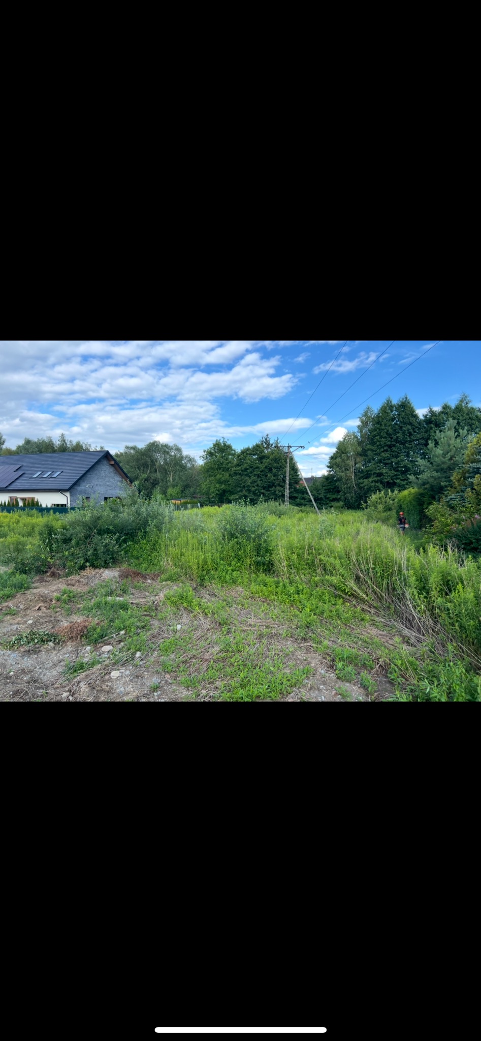 Zarośnięta działka z domem w tle; wysoka trawa i krzewy przed budynkiem. Widoczny słup energetyczny i częściowo osoba z narzędziem. Błękitne niebo z chmurami.