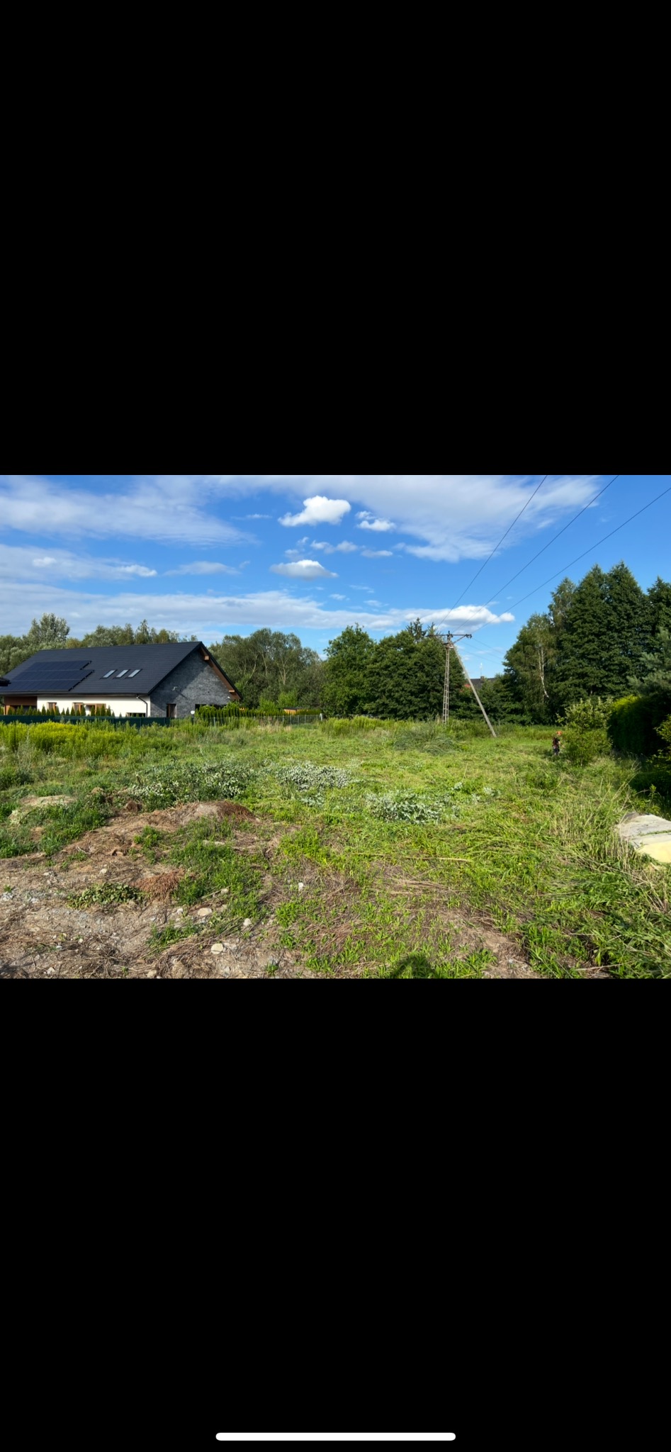 Działka z bujną trawą i fragmentem świeżo przekopanej ziemi. W tle dom z panelami słonecznymi i drzewa pod błękitnym niebem.