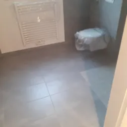 Szara, wykafelkowana łazienka z grzejnikiem drabinkowym i toaletą zabezpieczoną folią. Widok z perspektywy wejścia, po remoncie. Minimalistyczny design.