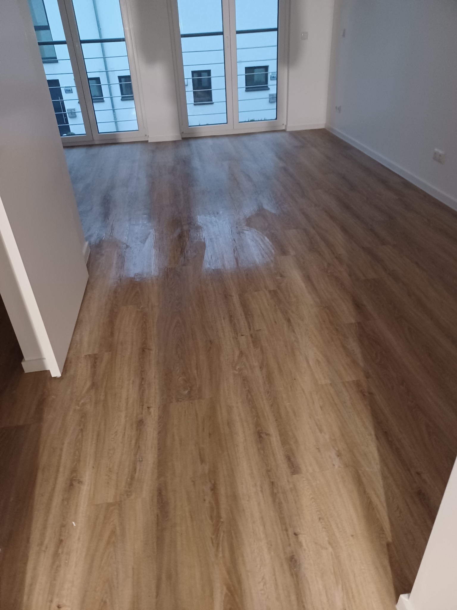 Ukończona podłoga z paneli drewnopodobnych w jasnym, nowoczesnym wnętrzu. Widoczne duże okna balkonowe i białe ściany. Parkiet ułożony równolegle do okien.