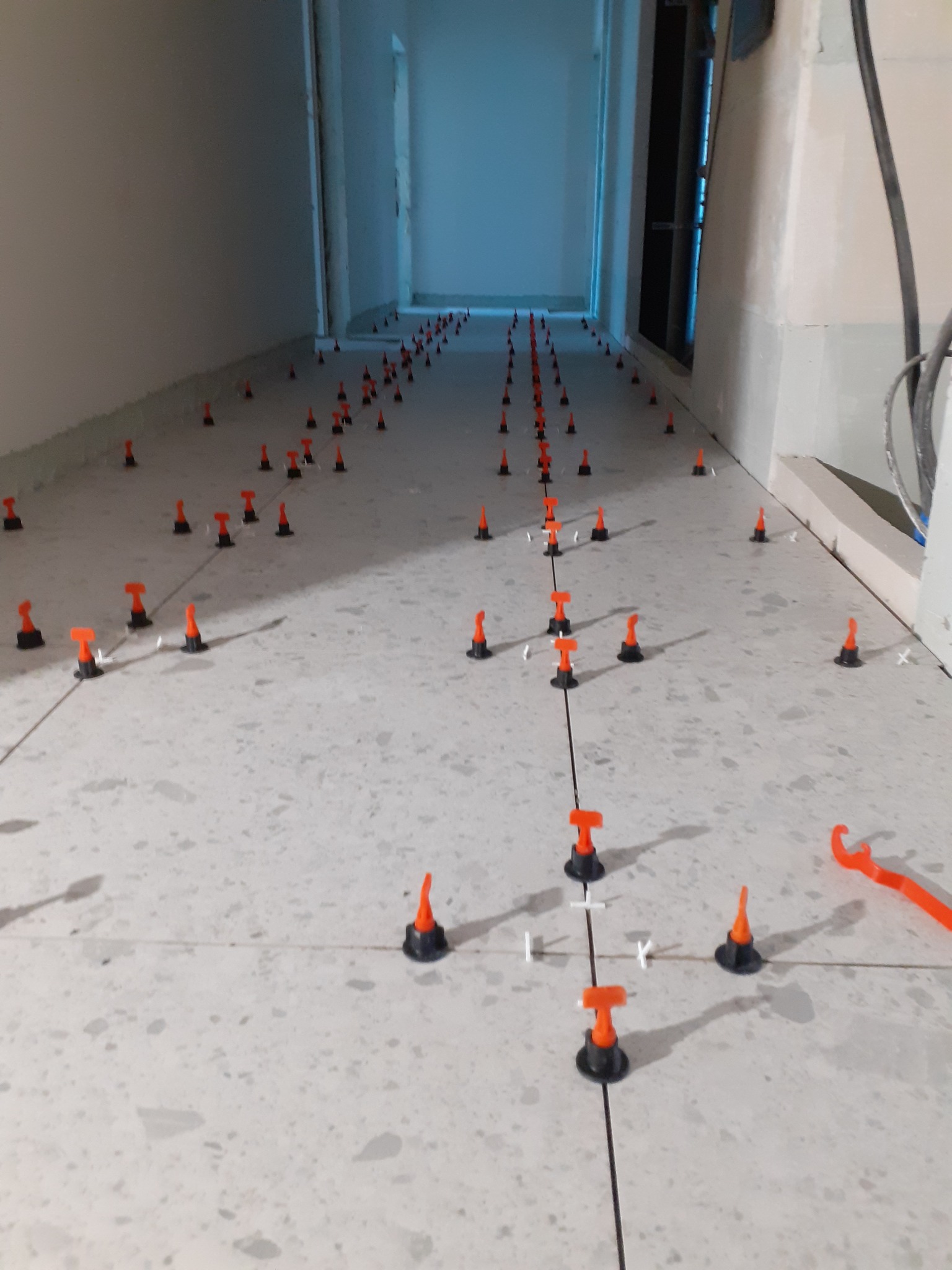 Układanie płytek na podłodze z użyciem systemu poziomowania, widoczne krzyżyki dystansowe i klipsy. Perspektywa w głąb korytarza, jasne płytki imitujące kamień.