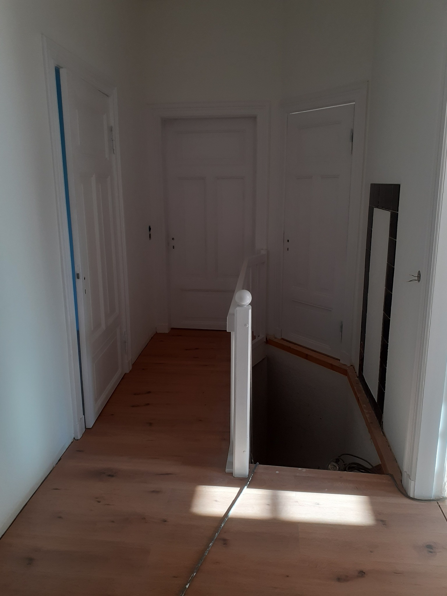 Jasne, drewniane panele podłogowe w korytarzu z białymi drzwiami. Widoczny fragment schodów do piwnicy z barierką. W tle drzwi i fragment ściany z czarno-białymi płytkami.