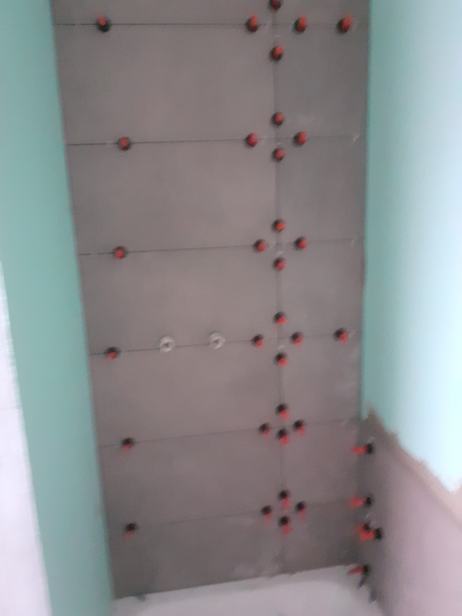 Ściana w trakcie układania szarych płytek z użyciem czerwonych krzyżyków dystansowych. Widoczne fragmenty zielonej ściany i biała instalacja.