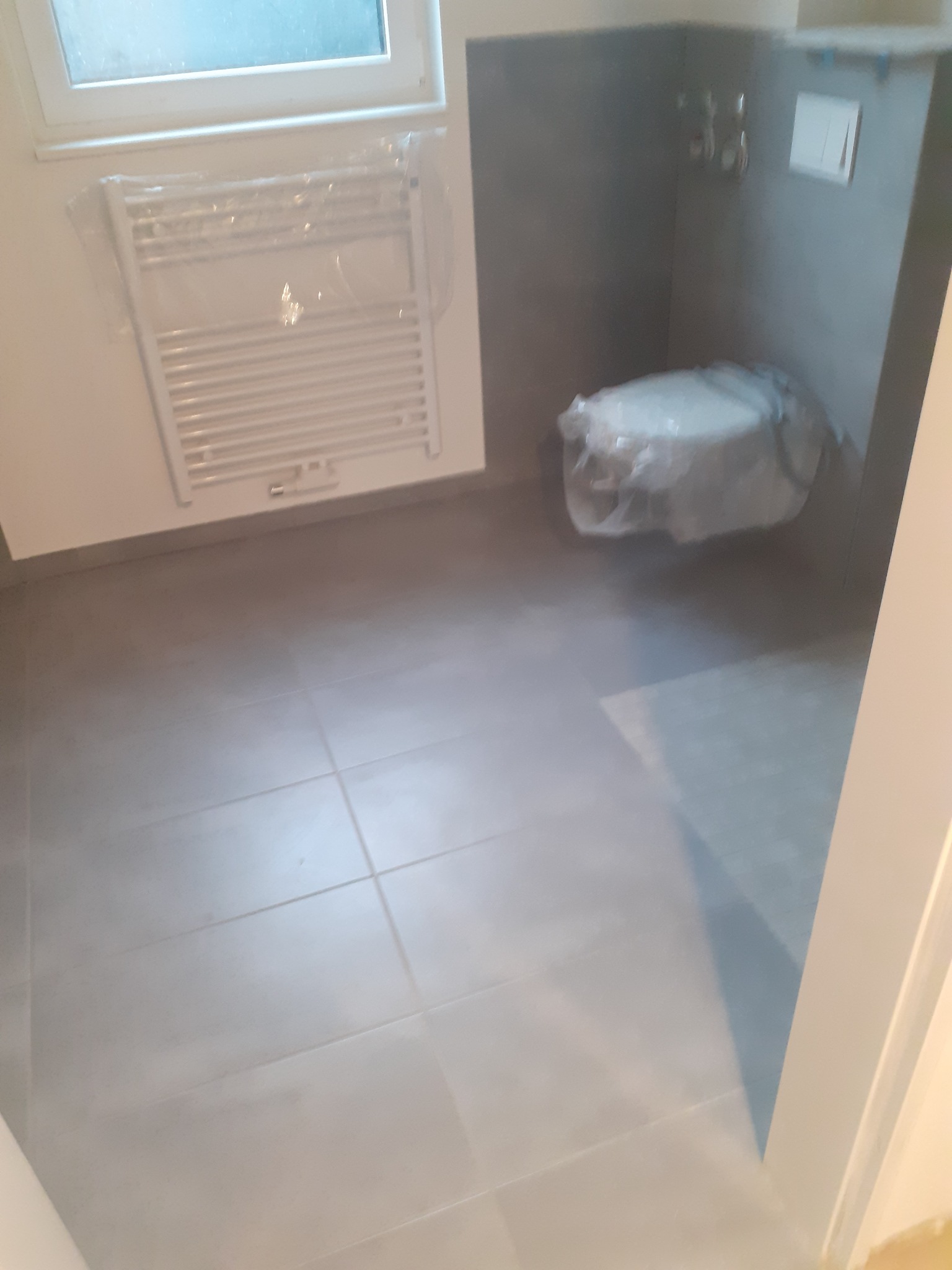 Szara, wykafelkowana łazienka z grzejnikiem drabinkowym i toaletą zabezpieczoną folią. Widok z perspektywy wejścia, po remoncie. Minimalistyczny design.