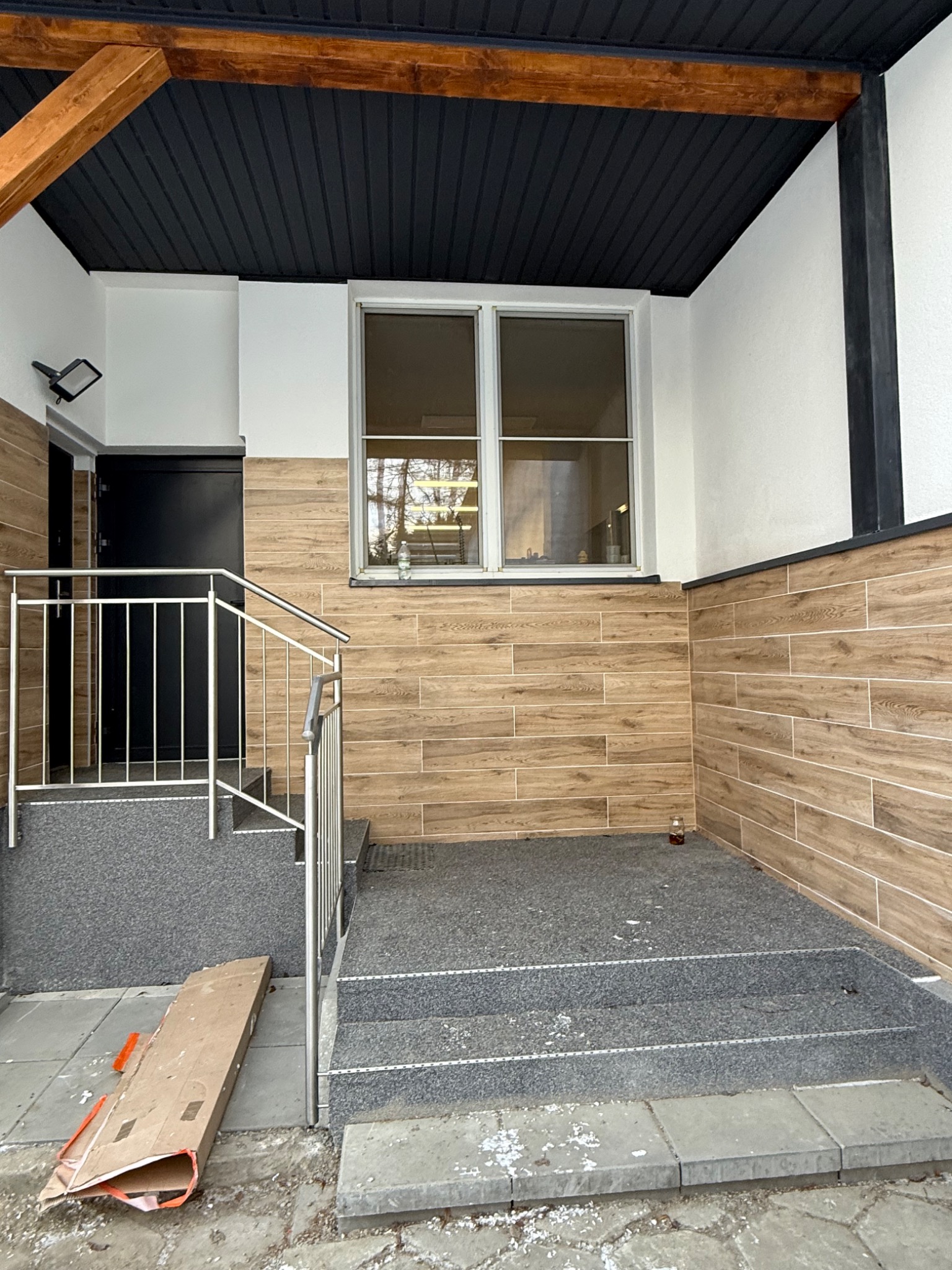 Wykończone wejście do budynku z drewnopodobną okładziną ścienną, szarymi schodami i metalową barierką. Widoczne okno i czarne drzwi wejściowe.