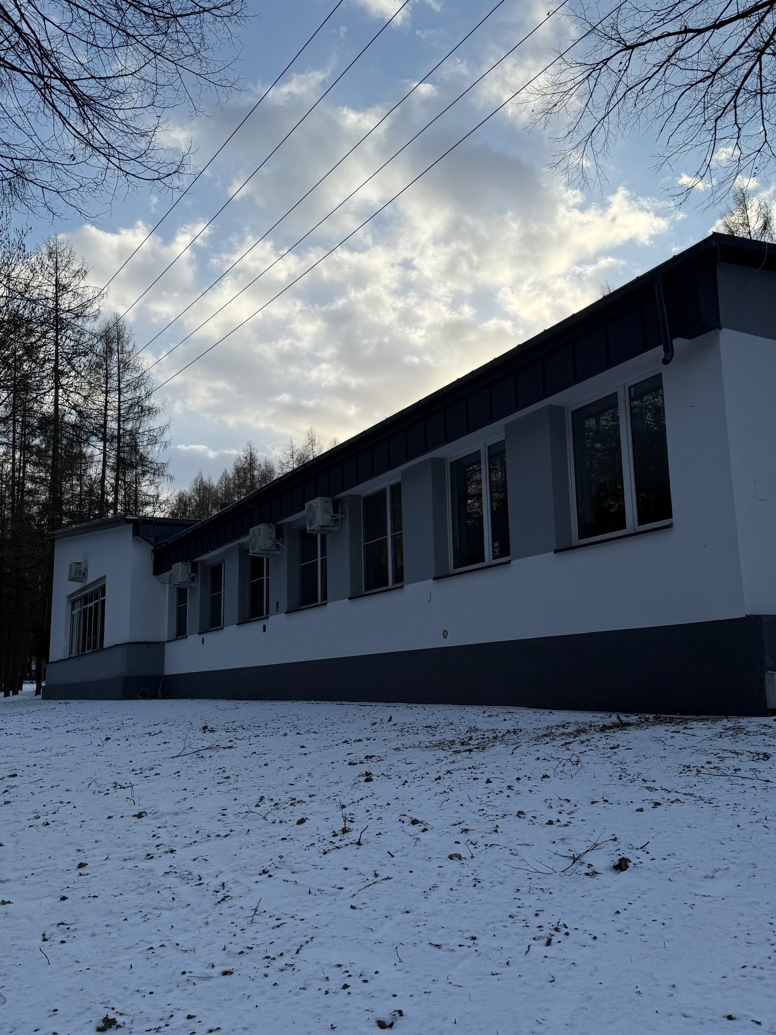 Biały budynek z klimatyzatorami na elewacji, widziany z dołu na tle zimowego krajobrazu i pochmurnego nieba z przewodami elektrycznymi.