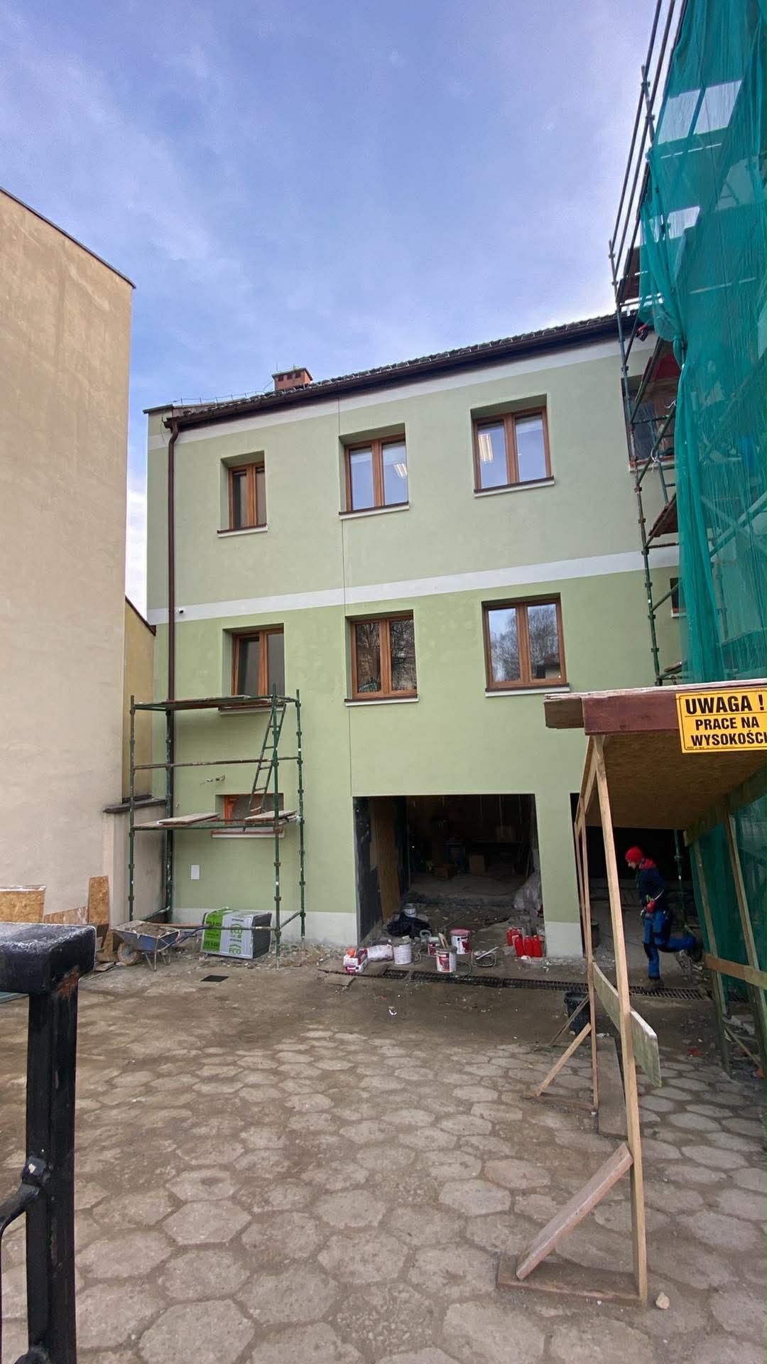 Dwukondygnacyjny budynek w trakcie malowania elewacji na zielono, z widocznymi rusztowaniami i narzędziami malarskimi. Tablica ostrzegawcza o pracy na wysokości.