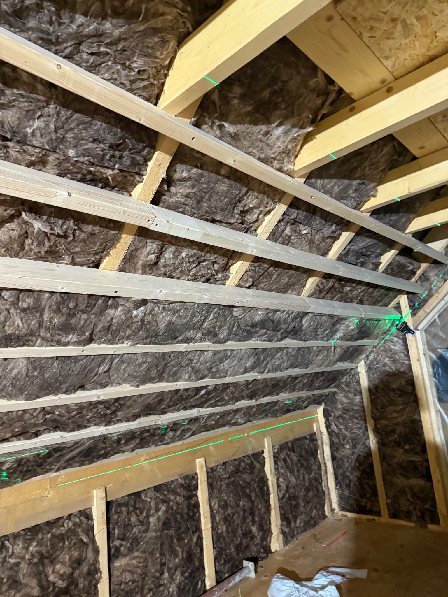 Skośny kadr na ocieplone wełną mineralną poddasze z drewnianą konstrukcją. Widoczne zielone linie lasera wyznaczające poziom. Prace izolacyjne w toku.