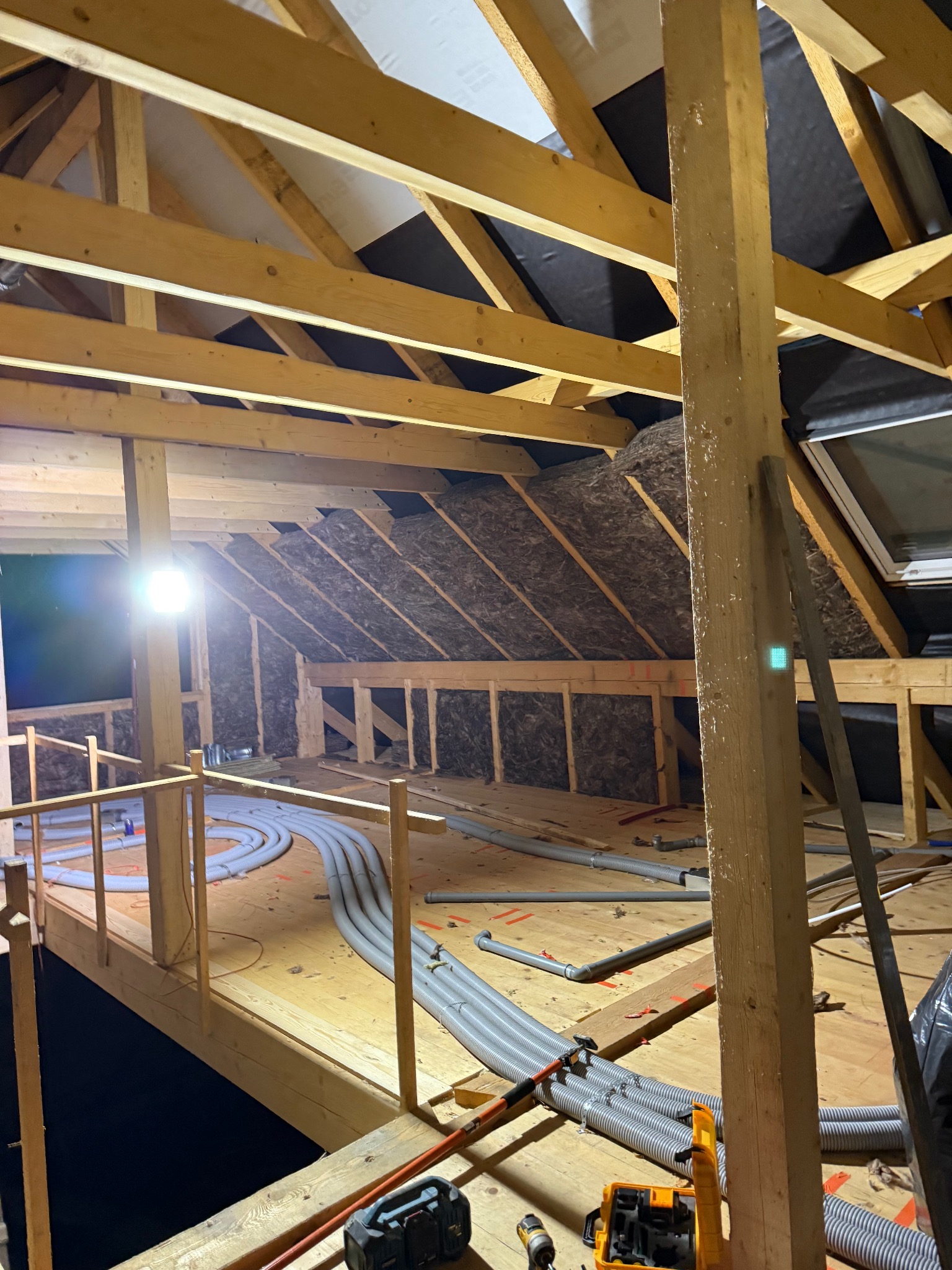 Poddasze w trakcie adaptacji: widoczne drewniane elementy konstrukcyjne, izolacja, okno dachowe i szare rury wentylacyjne. Oświetlenie robocze. Etap prac wykończeniowych.