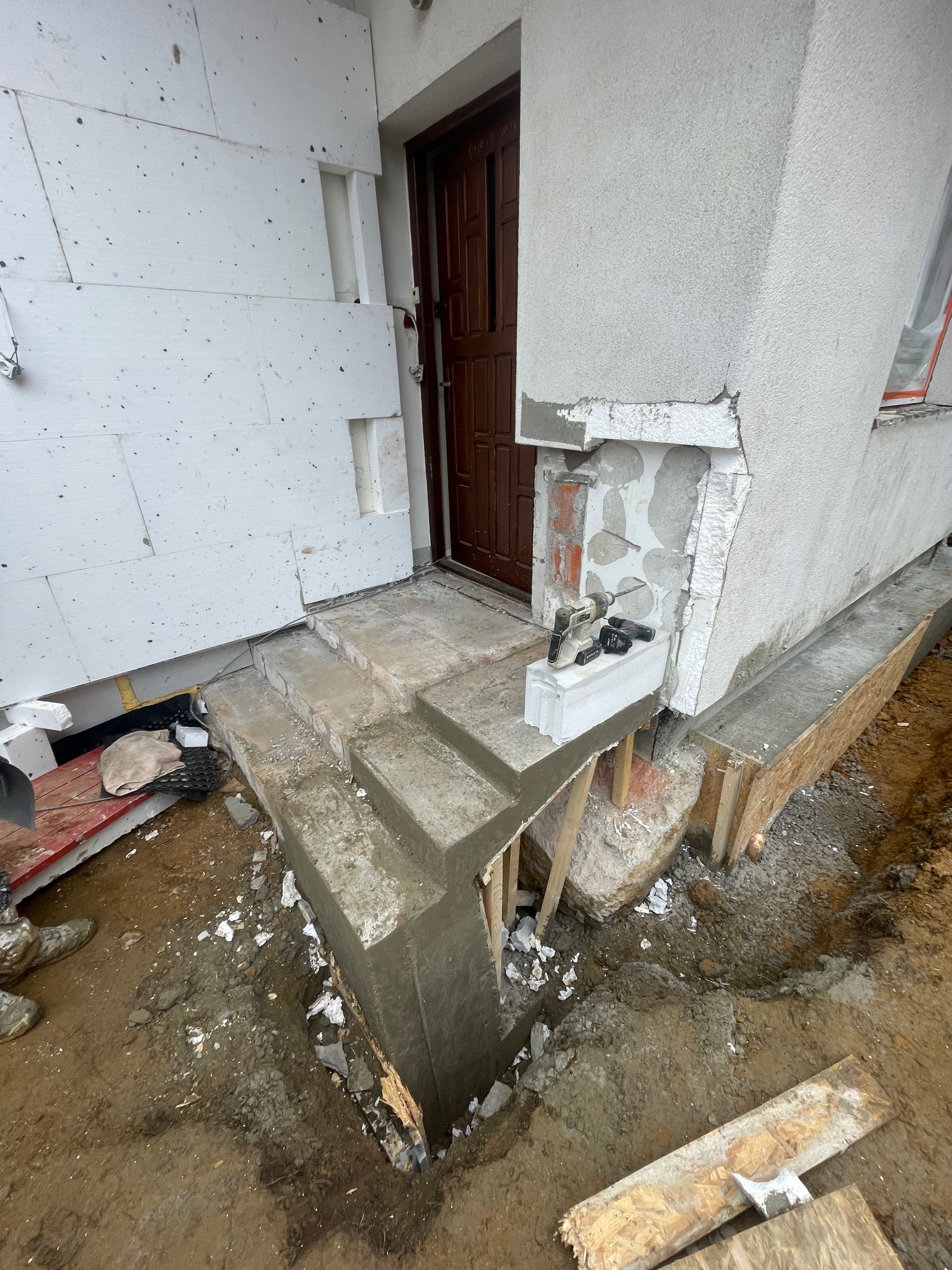 Budowa schodów wejściowych do domu z ociepleniem elewacji. Widoczne narzędzia i materiały budowlane. Etap prac w toku, z wykopem i częściowo wykończonymi schodami.