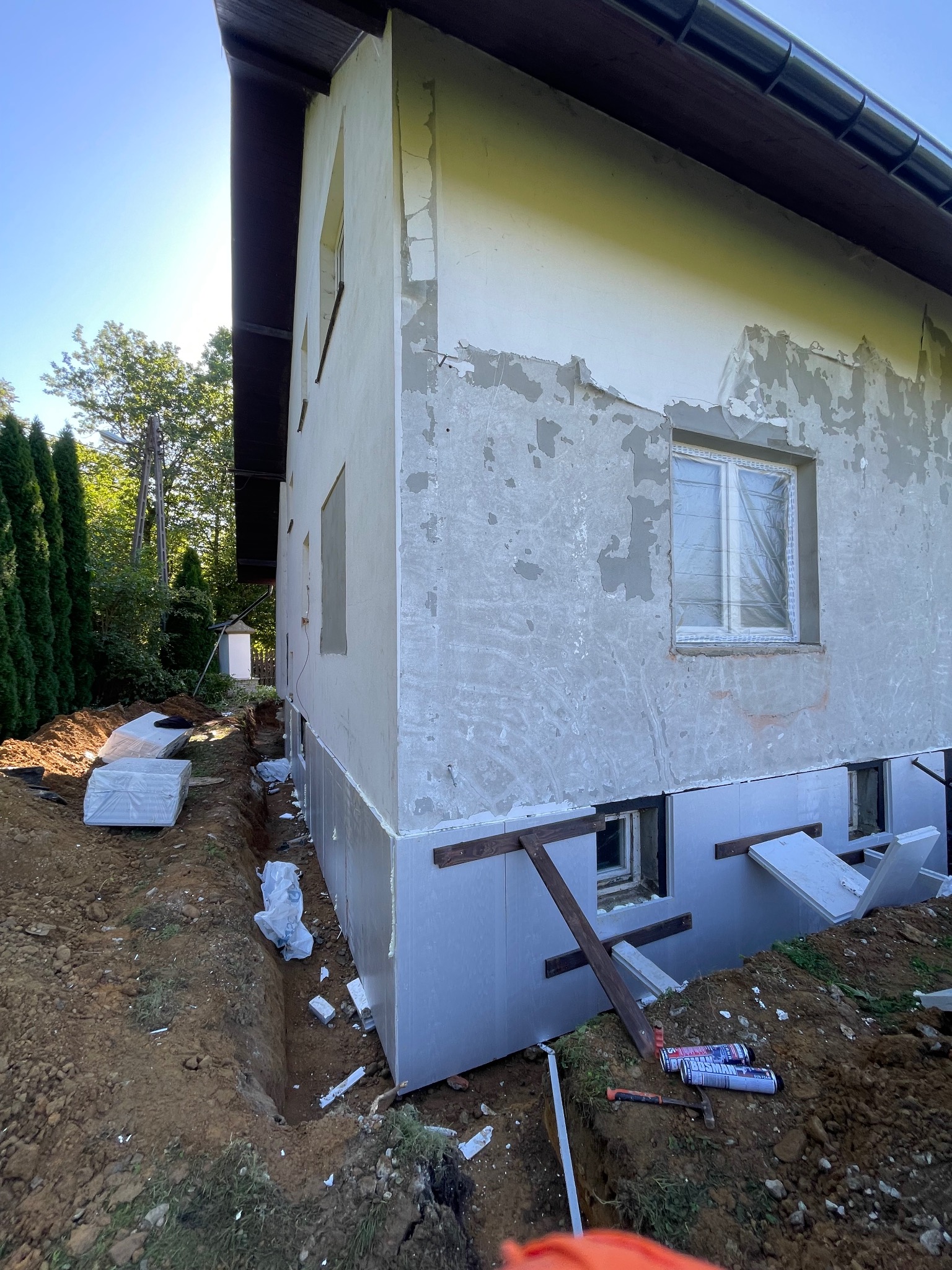 Ocieplanie fundamentów domu jednorodzinnego. Widoczne płyty izolacyjne, wykop i narzędzia. Częściowo otynkowana ściana budynku w tle.