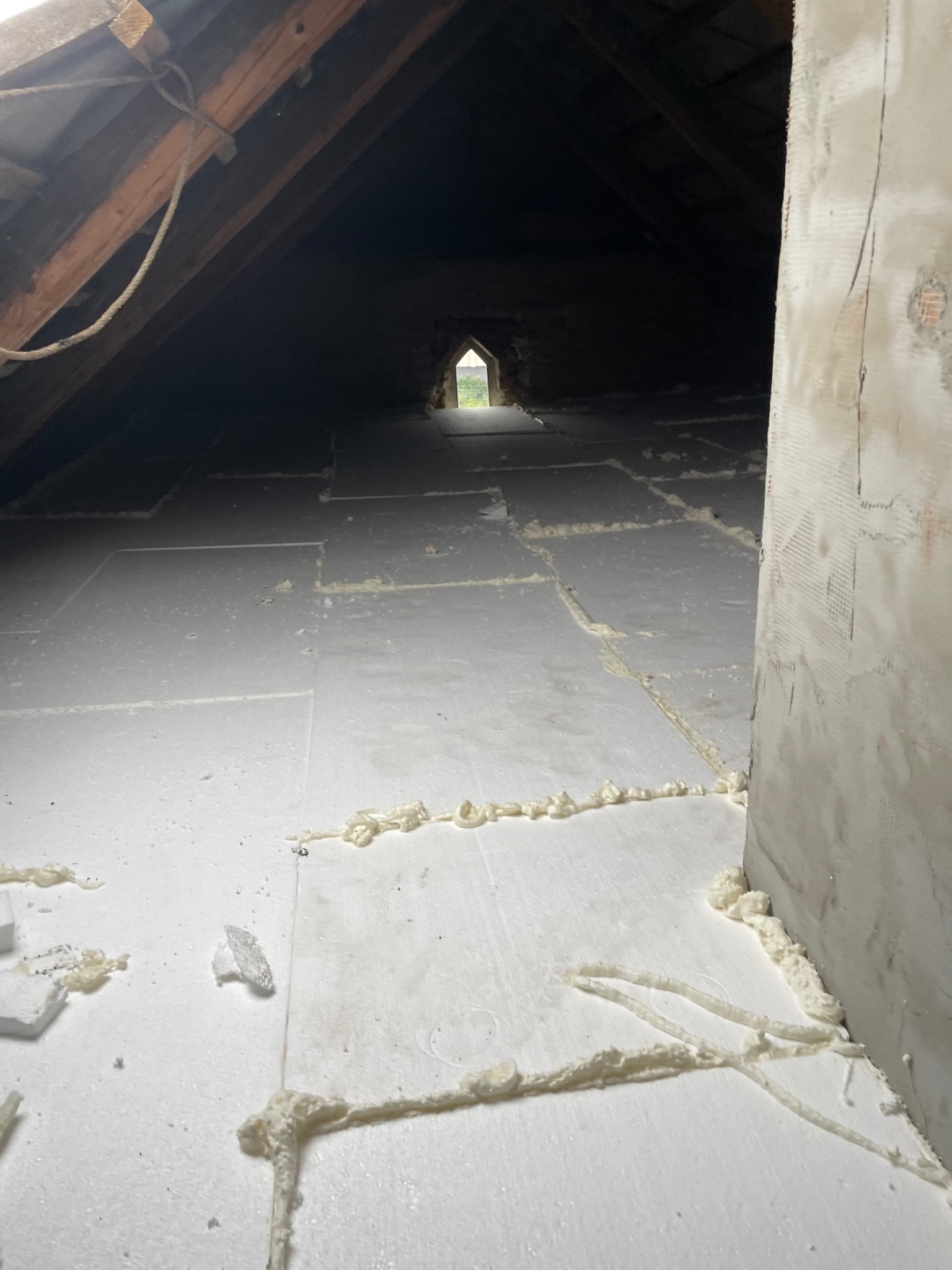 Poddasze z widoczną izolacją termiczną z białych płyt, uszczelnionych pianką montażową. W tle widoczne drewniane elementy konstrukcyjne dachu i okno.