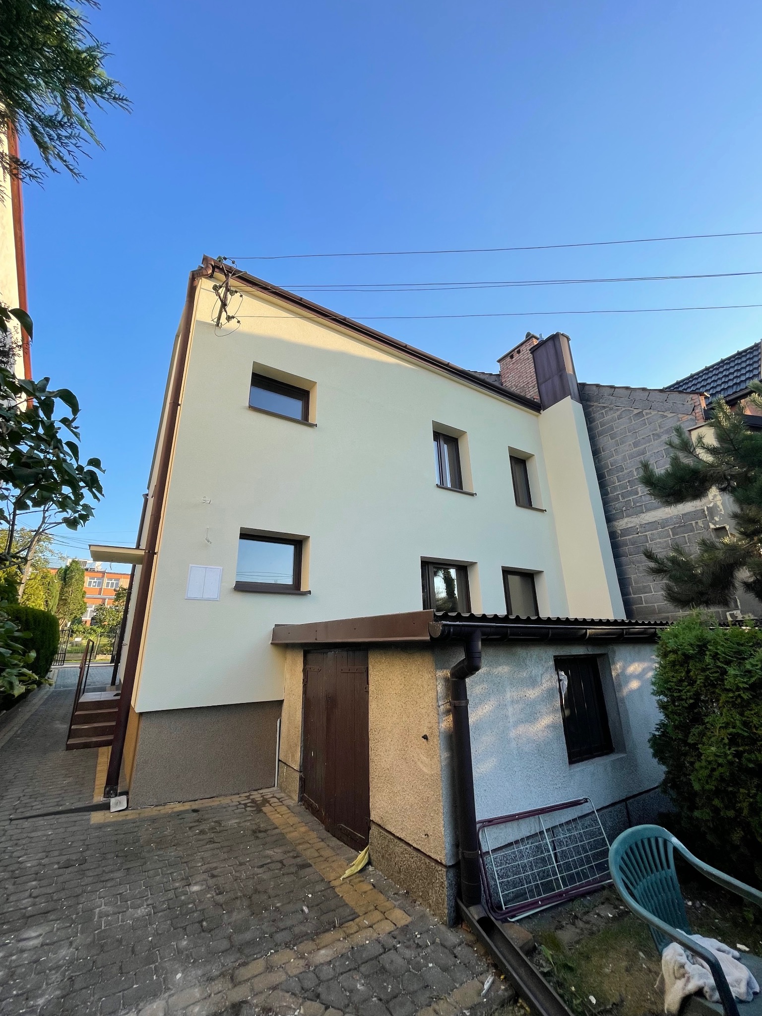 Dwupiętrowy dom z jasną elewacją, brązowymi ramami okiennymi i dachem, widziany z dołu pod kątem, z fragmentem chodnika z kostki brukowej i zieleni w tle.