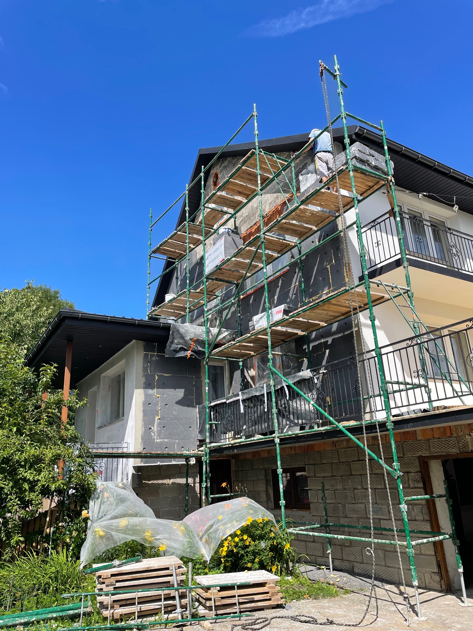 Dwukondygnacyjny dom w trakcie ocieplania elewacji, z widocznymi rusztowaniami i pracownikiem na górze. Część elewacji pokryta izolacją, a część jeszcze nieocieplona.