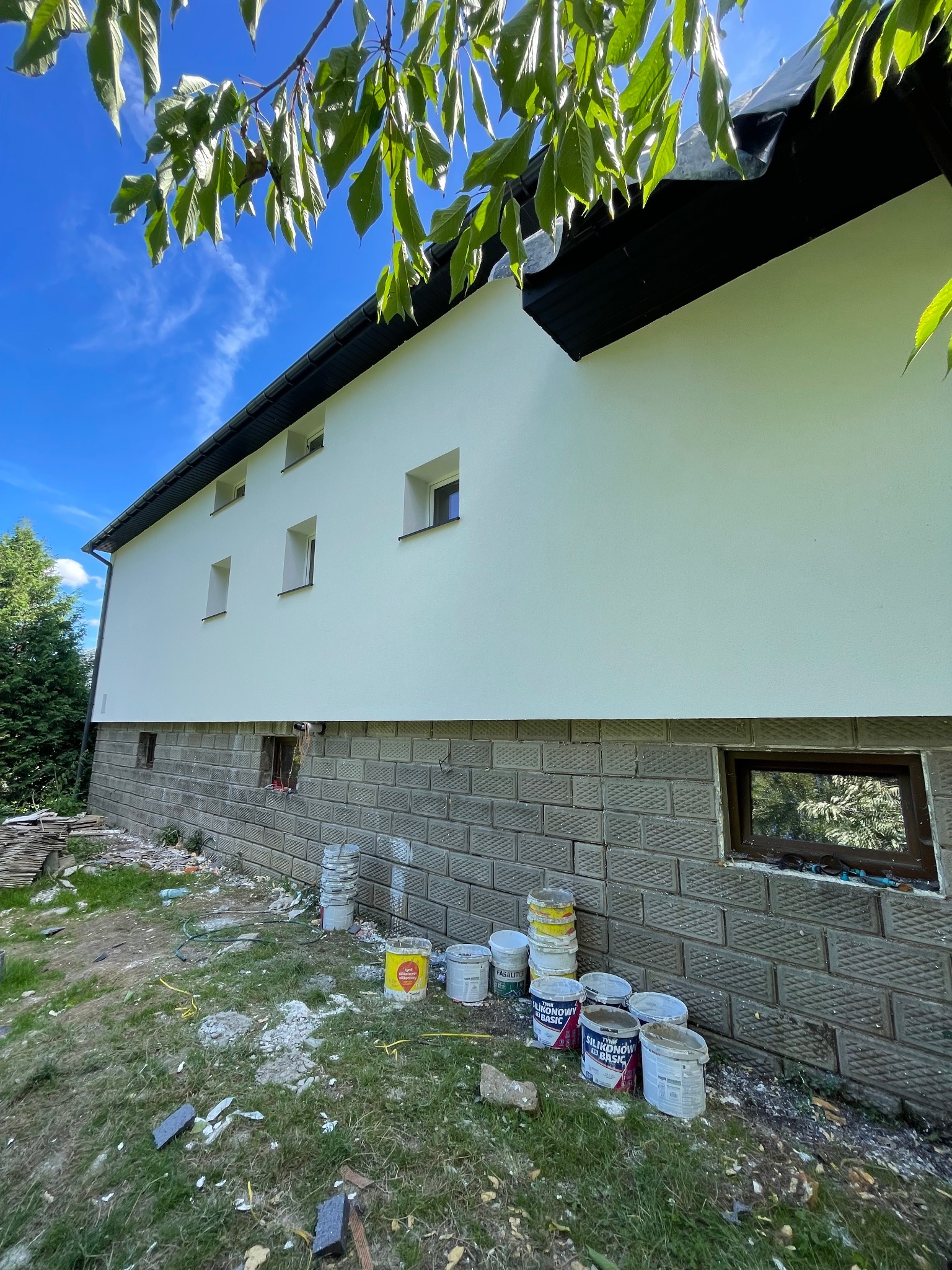 Ocieplona elewacja domu jednorodzinnego z widocznymi puszkami po farbie i resztkami materiałów budowlanych wokół, sugerującymi trwające prace wykończeniowe.