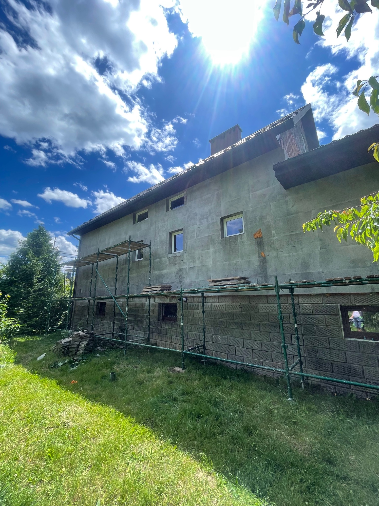 Dwukondygnacyjny dom w trakcie prac elewacyjnych, z widocznymi rusztowaniami i fragmentami niezabezpieczonej ściany. Słoneczny dzień, błękitne niebo z chmurami w tle.