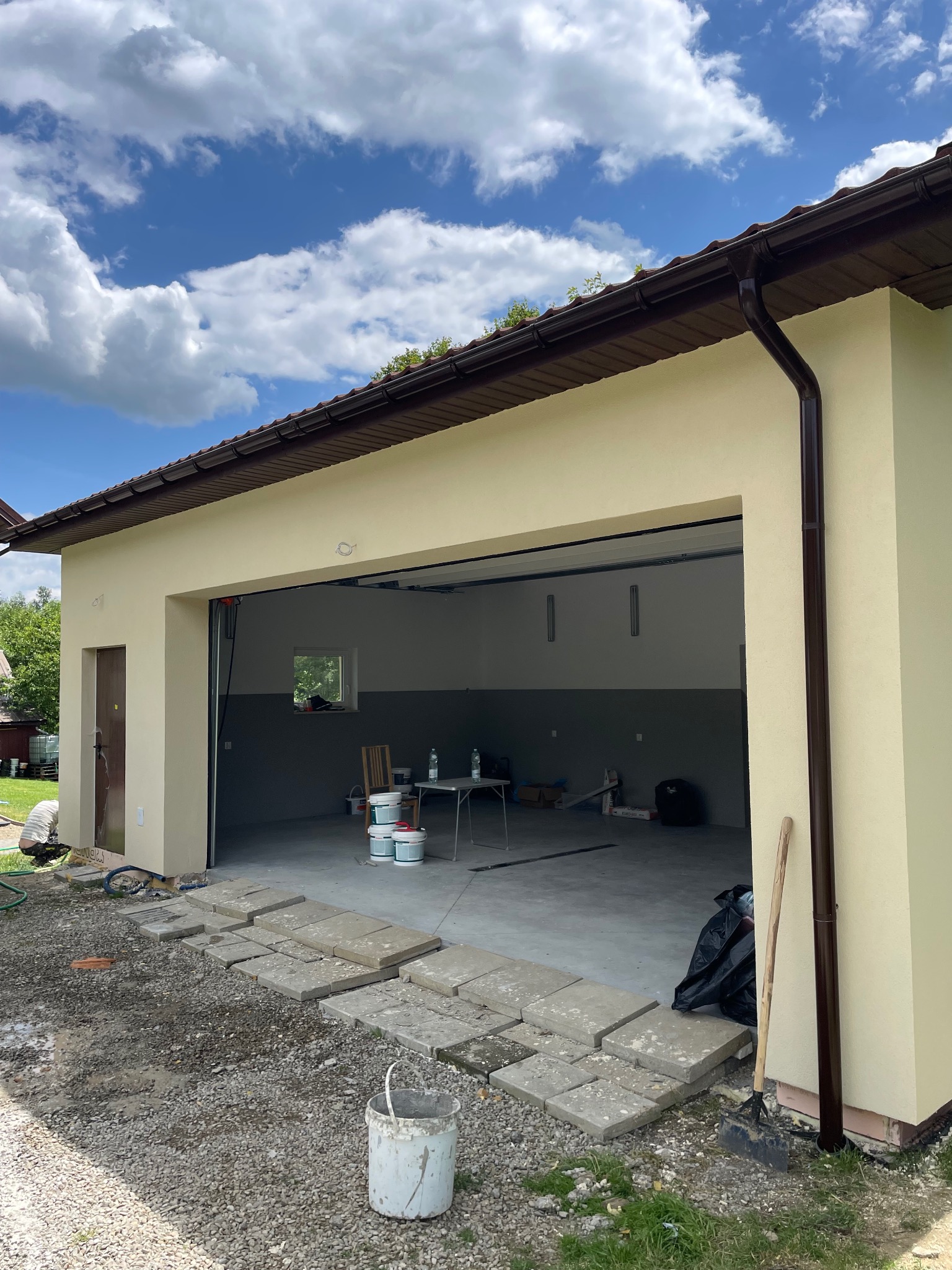 Otwarty garaż z beżową elewacją i brązowymi rynnami. Wnętrze szare, widoczne narzędzia i farby. Na zewnątrz ułożone płyty chodnikowe i wiadro.