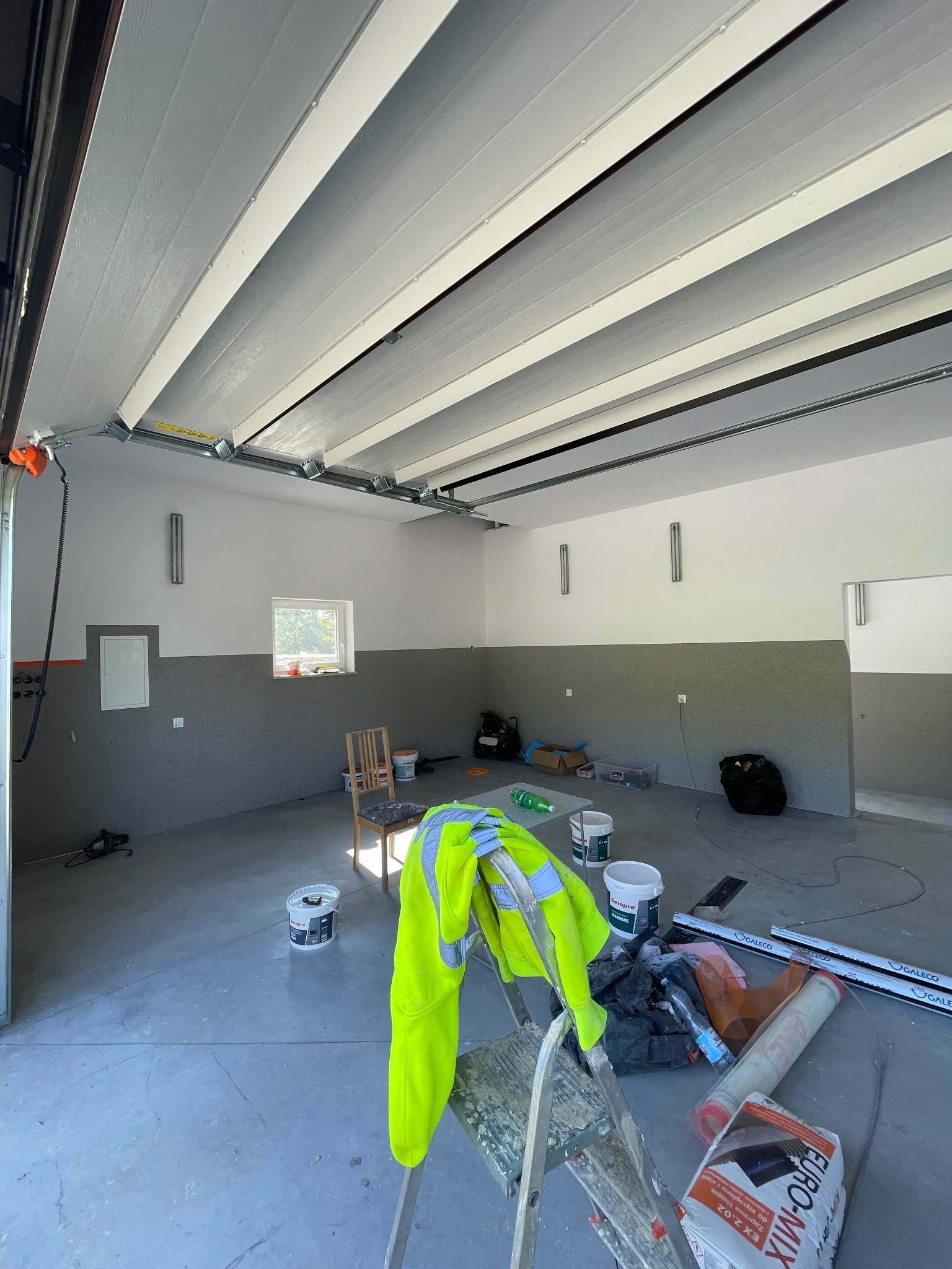 Wnętrze garażu po malowaniu ścian na szaro i biało, widoczne puszki z farbą, drabina z odblaskową kurtką i materiały budowlane. Świeżo odnowione pomieszczenie.