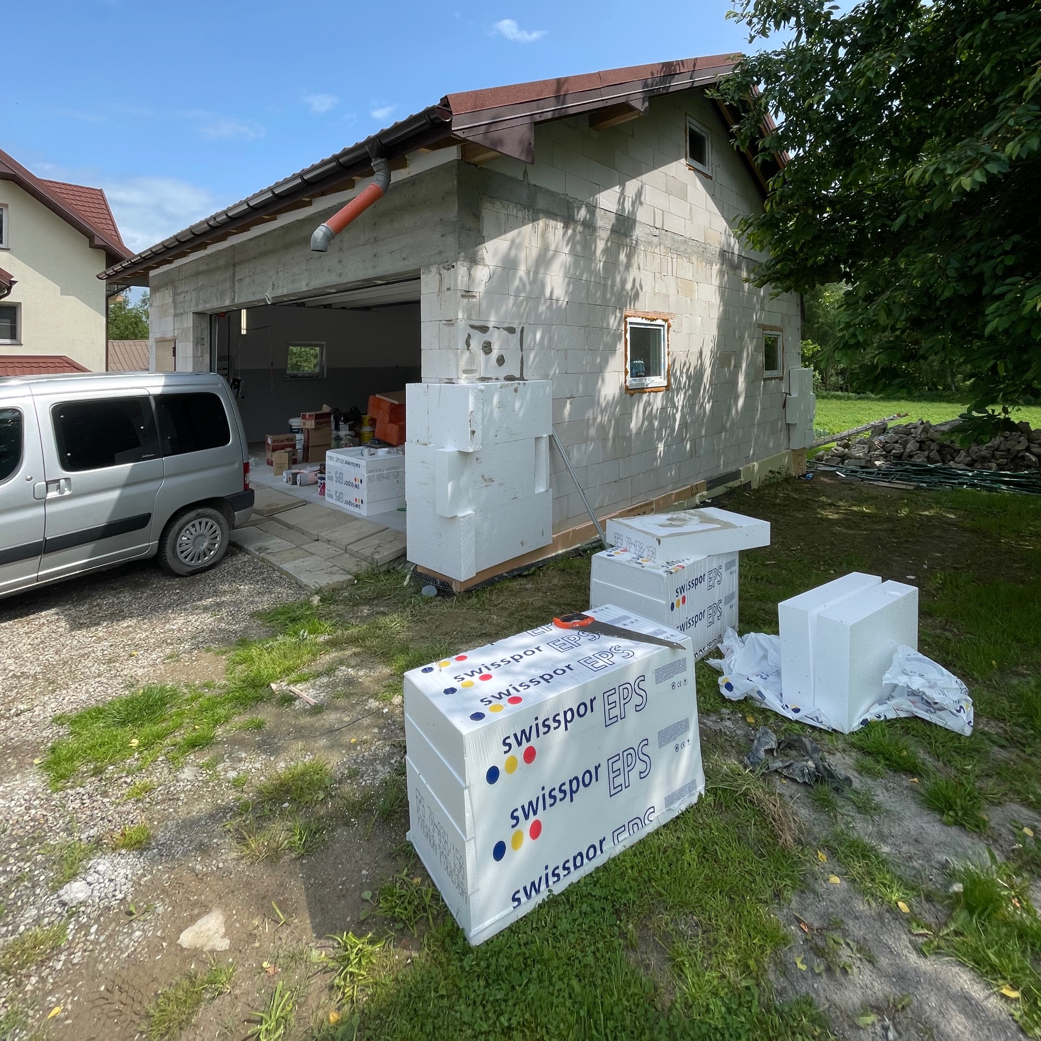Budynek w trakcie ocieplania, widoczne bloki styropianu Swissopor EPS, częściowo ocieplona ściana, okno i samochód dostawczy w tle. Prace budowlane w słoneczny dzień.
