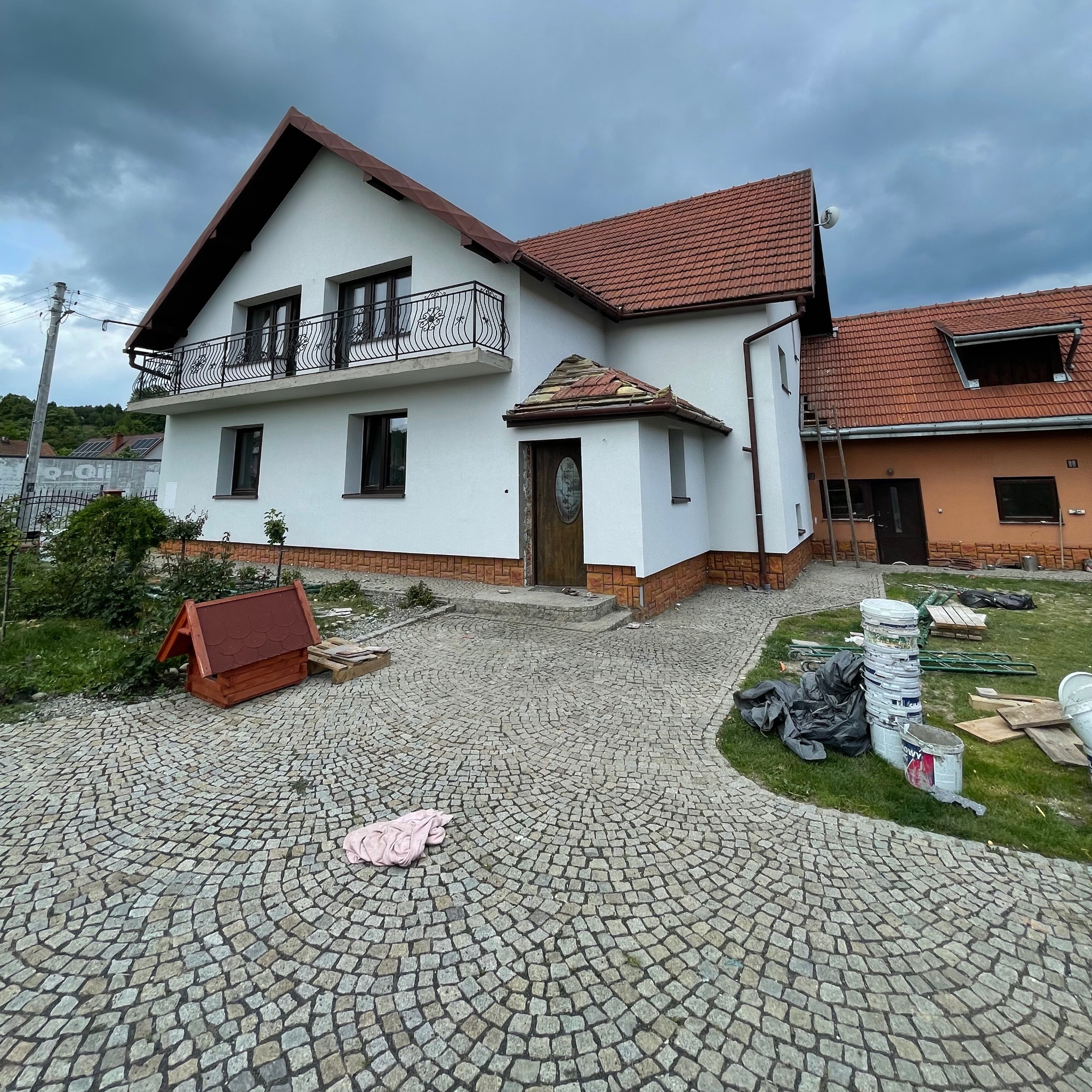 Biały dom z balkonem i czerwoną dachówką, widok z brukowanego podjazdu. Widoczne farby i akcesoria malarskie sugerują prace wykończeniowe.
