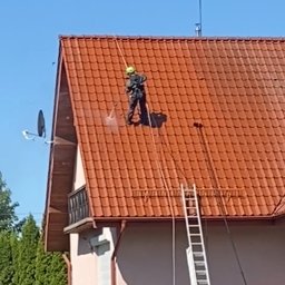 P.H.U TOLA - Pracownik w kasku i kombinezonie, zabezpieczony linami, czyści dach z czerwonej dachówki za pomocą myjki ciśnieniowej. Obok drabina i fragment elewacji domu.