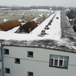 P.H.U TOLA - Zimowy widok z góry na rozległy kompleks budynków z płaskimi dachami pokrytymi śniegiem, w tle widoczne drzewa i zabudowa miejska w pochmurny dzień.