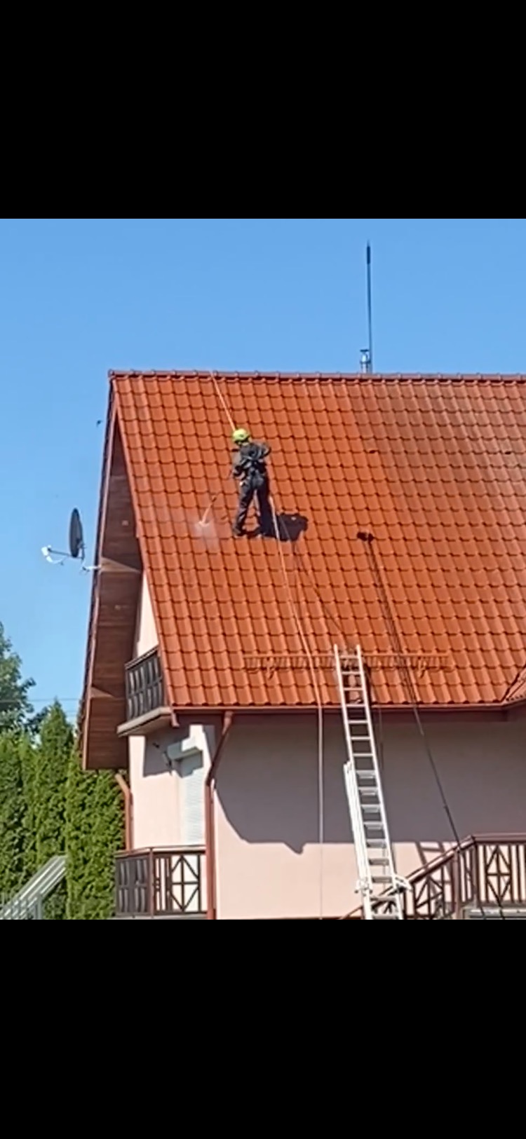 Pracownik w kasku i kombinezonie, zabezpieczony linami, czyści dach z czerwonej dachówki za pomocą myjki ciśnieniowej. Obok drabina i fragment elewacji domu.