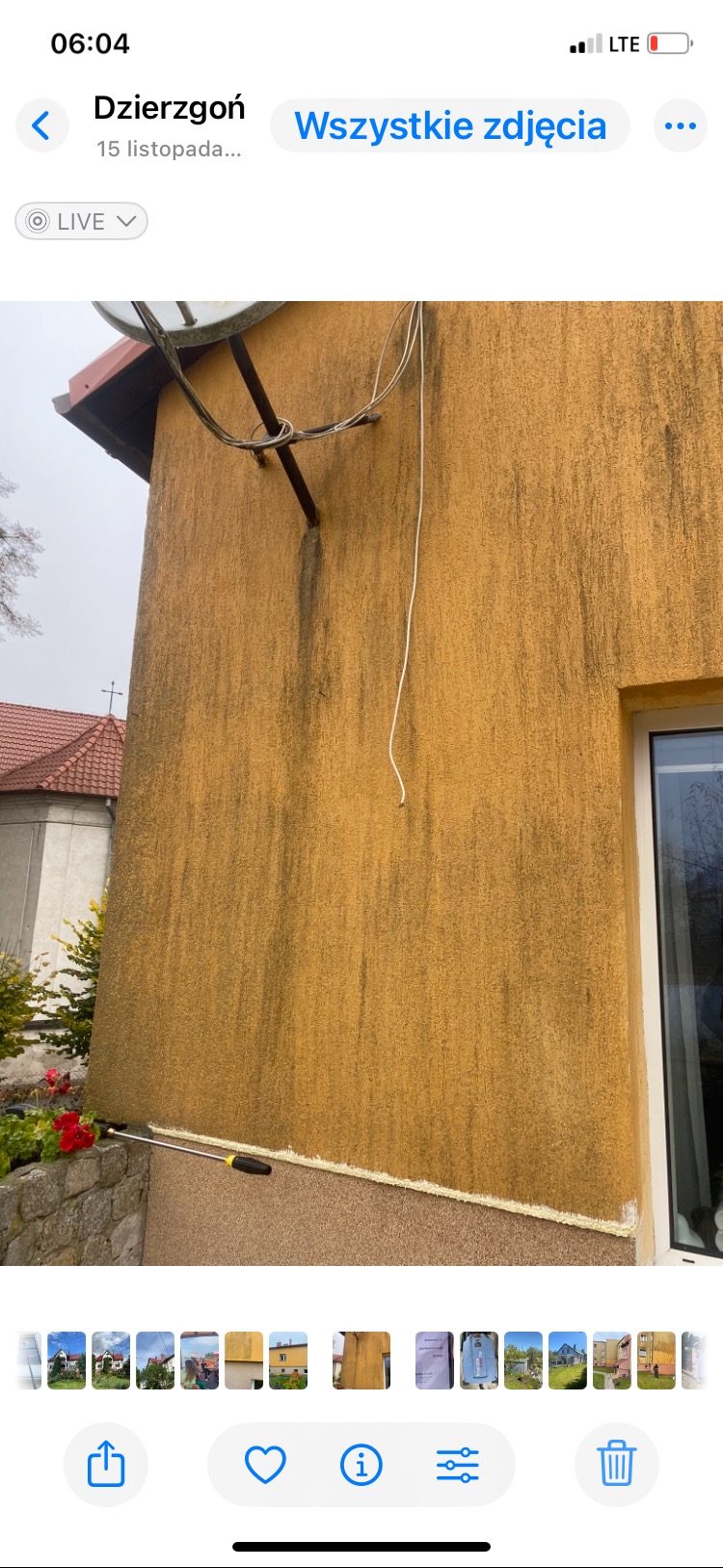 Żółta elewacja z widocznymi zabrudzeniami i zaciekami, częściowo umyta. W tle fragment budynku z czerwoną dachówką i okno. Widoczny sprzęt do mycia elewacji.