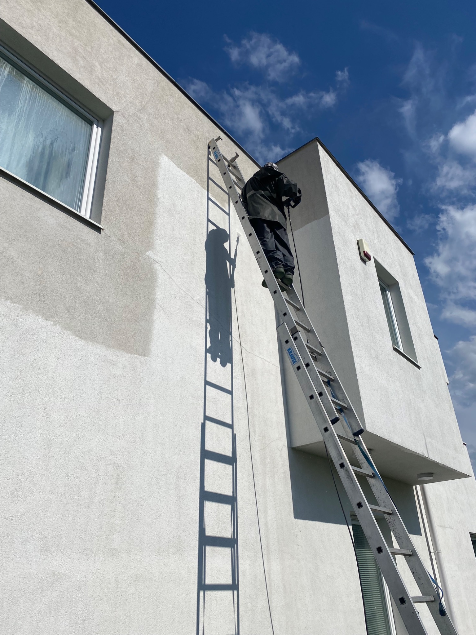 Pracownik na drabinie maluje fragment elewacji nowoczesnego domu. Widoczny świeżo pomalowany obszar, kontrastujący z resztą ściany. Błękitne niebo w tle.