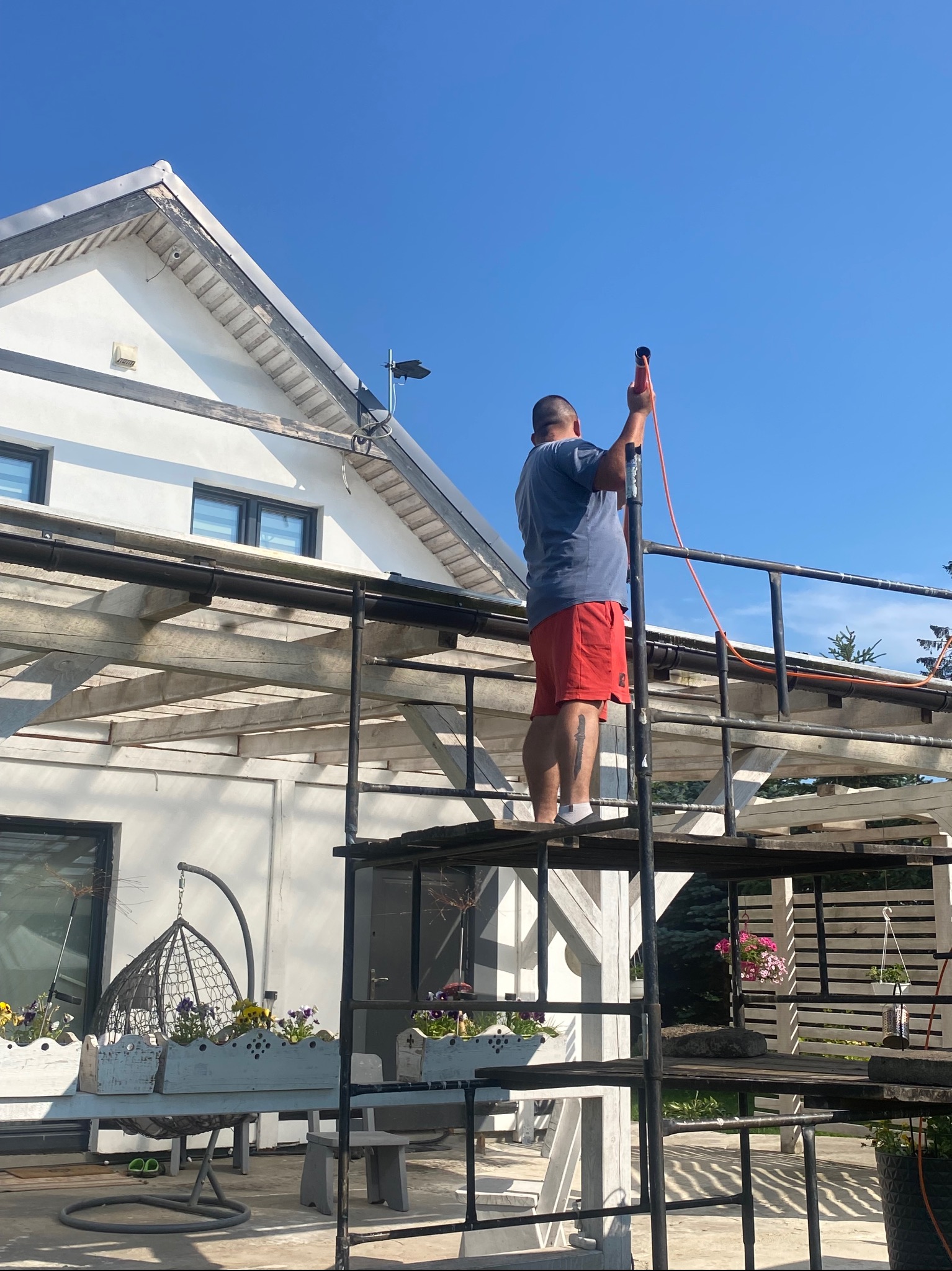 Pracownik na rusztowaniu z lancą ciśnieniową przygotowuje się do pracy przy elewacji domu z białym tynkiem i drewnianymi elementami wykończeniowymi.