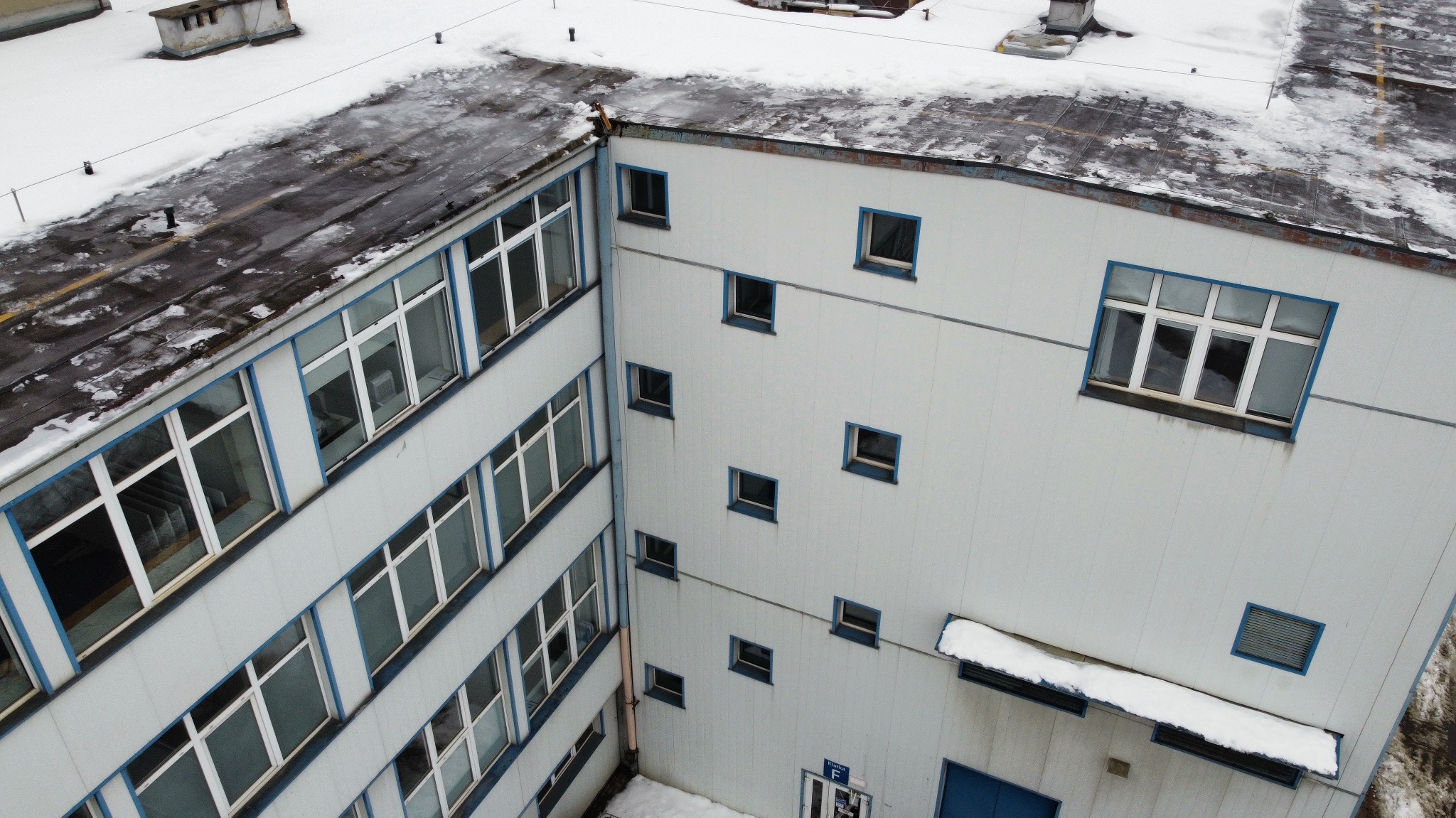 Wysoki budynek przemysłowy z płaskim dachem pokrytym śniegiem, widoczny z góry. Biała elewacja z niebieskimi ramami okiennymi. Częściowo ośnieżony teren wokół budynku.