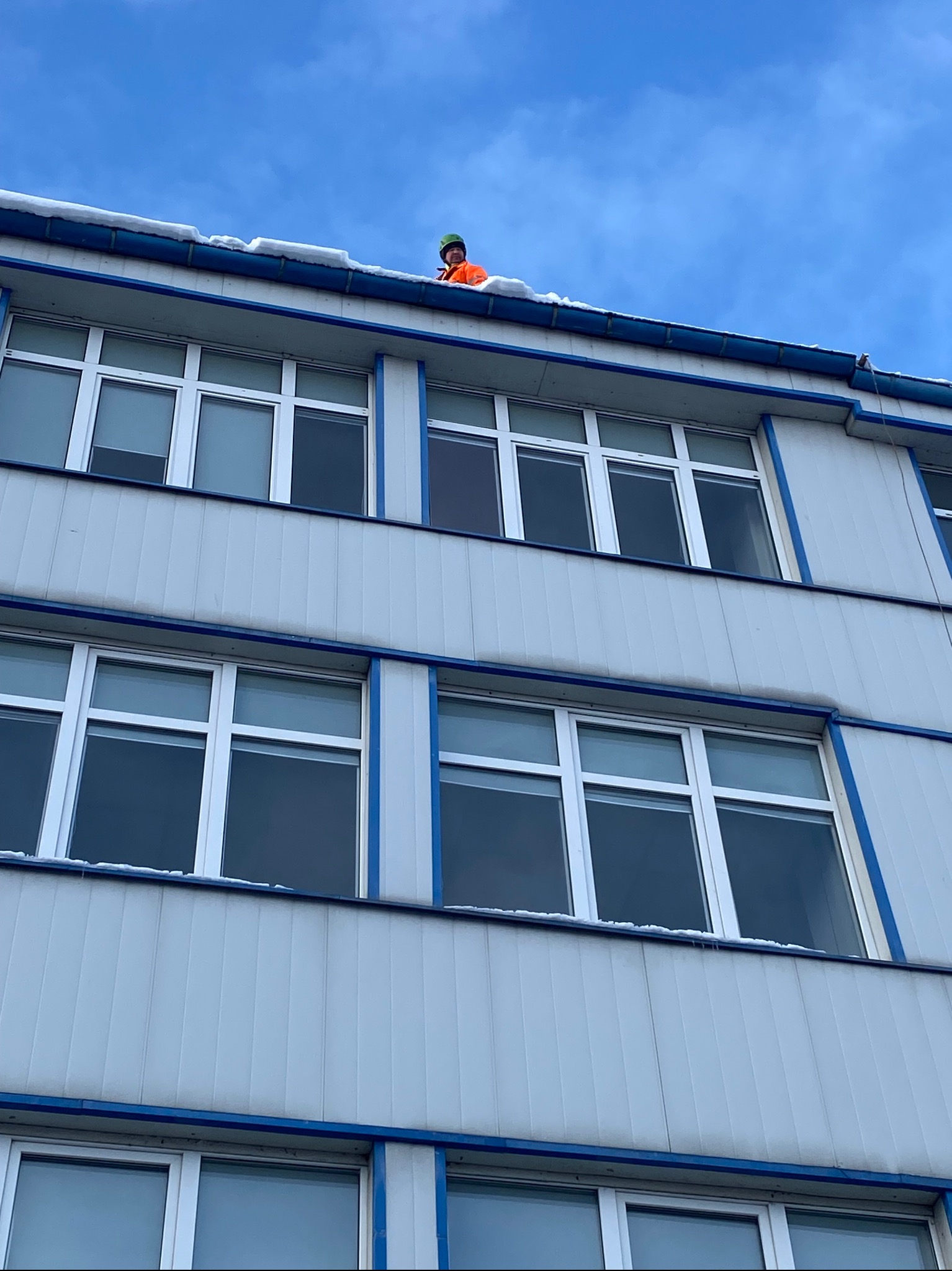 Pracownik w pomarańczowym kombinezonie usuwa śnieg z dachu budynku z szarej elewacji z niebieskimi akcentami i rzędami okien, widok z dołu na błękitne niebo.