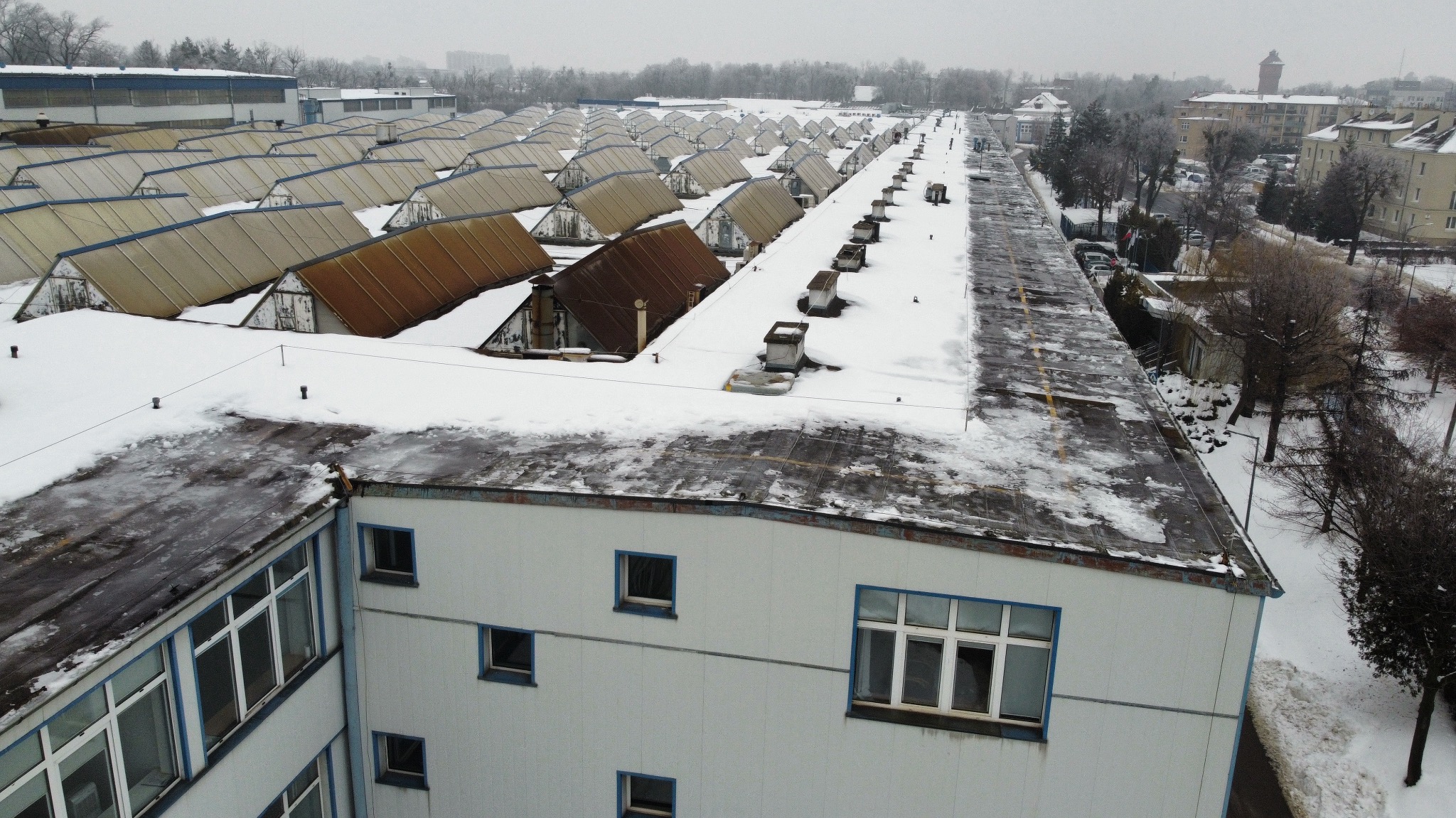 Zimowy widok z góry na rozległy kompleks budynków z płaskimi dachami pokrytymi śniegiem, w tle widoczne drzewa i zabudowa miejska w pochmurny dzień.