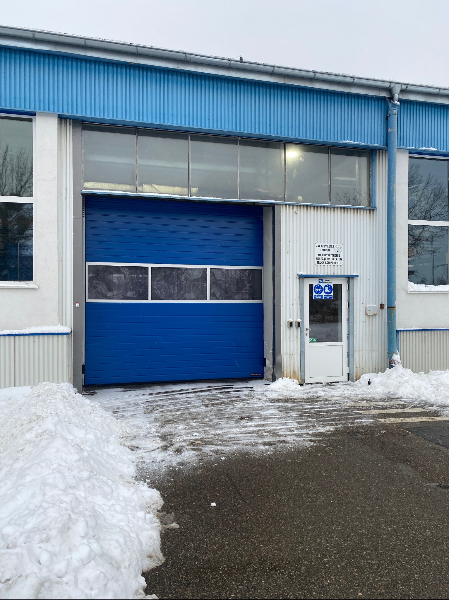 Błękitna brama segmentowa z oknami w hali przemysłowej, z fragmentami śniegu na podjeździe i dachu. Widoczny fragment elewacji z drzwiami i tabliczkami informacyjnymi.