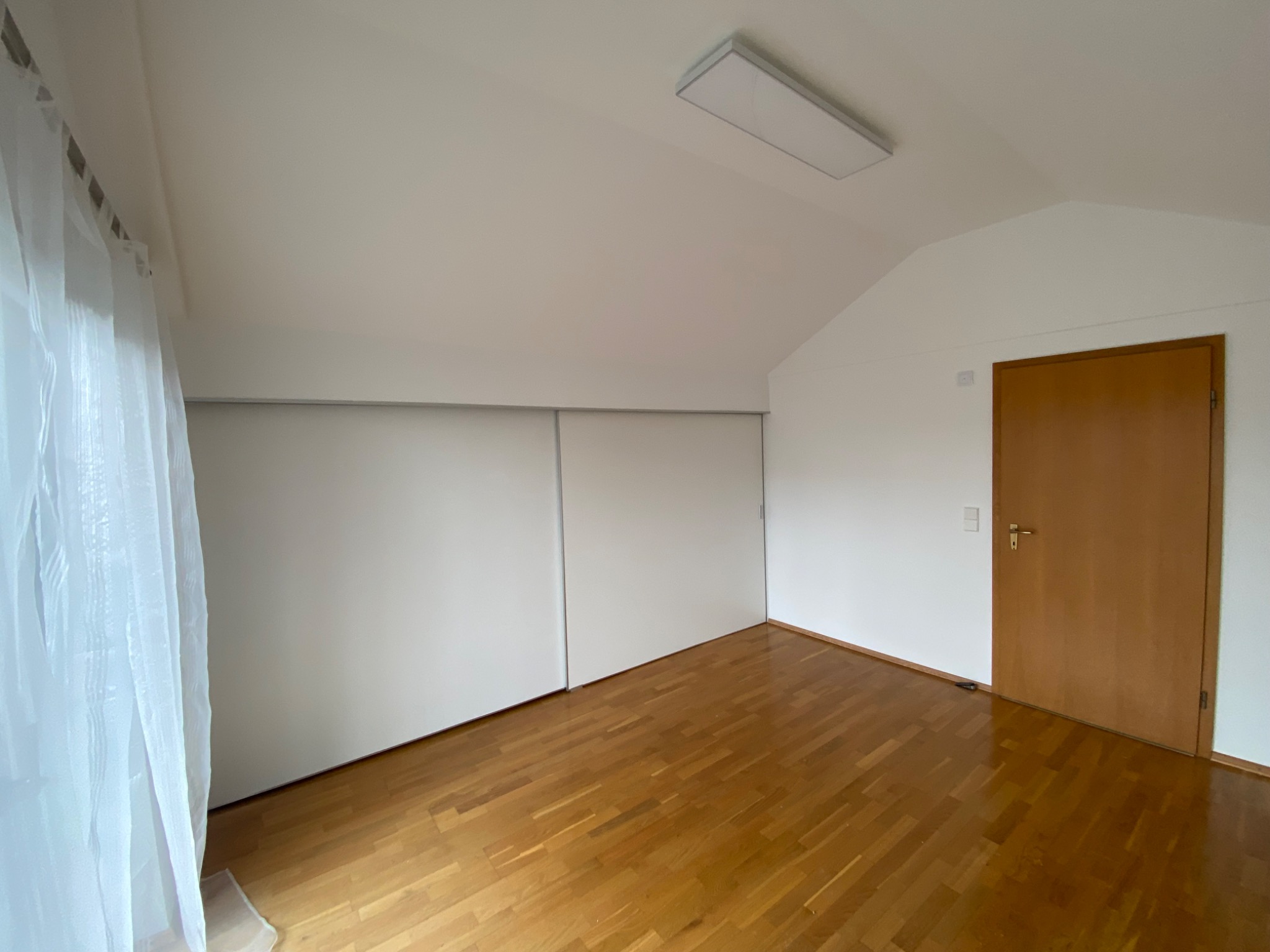 Jasne wnętrze poddasza z drewnianą podłogą, białymi ścianami i przesuwnymi drzwiami szafy. Widoczne okno z firanką i drewniane drzwi. Minimalistyczny design.