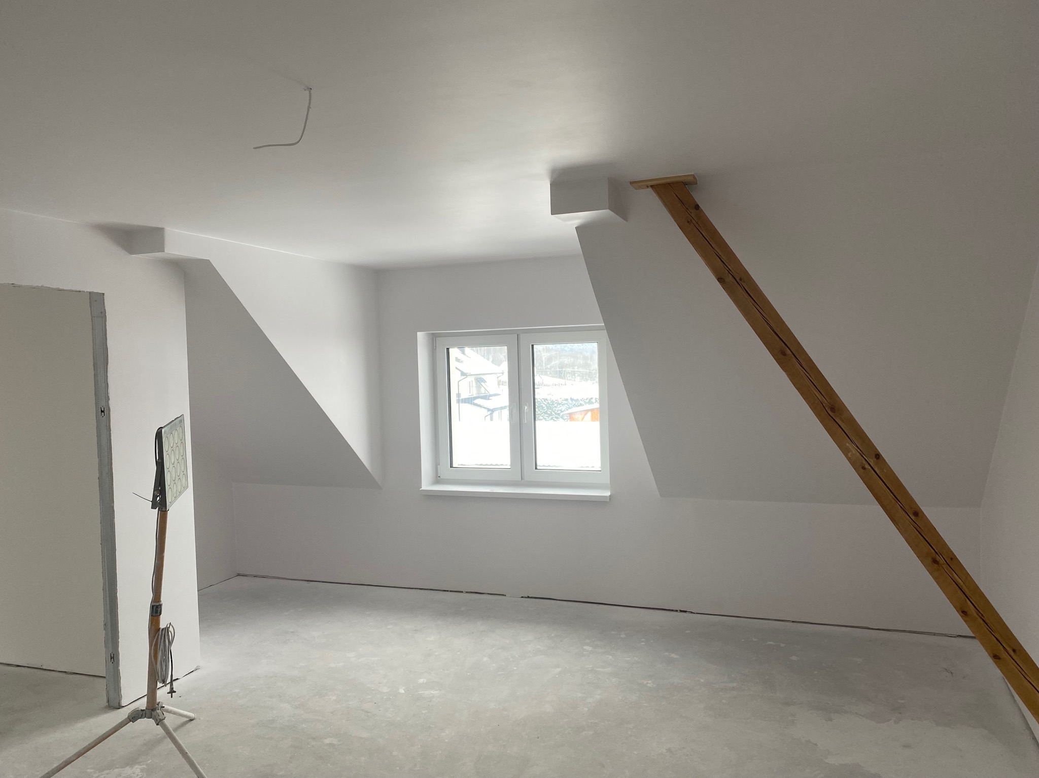Białe poddasze po szpachlowaniu, z oknem i drewnianą podporą. Widoczna lampa na statywie. Świeżo wykończone ściany i sufit, surowa podłoga. Minimalistyczny, jasny kadr.