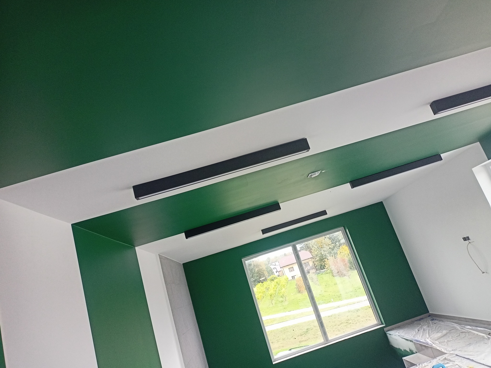 Pokój po malowaniu: zielone ściany i sufit z białymi pasami, czarne lampy sufitowe, okno z widokiem na zewnątrz. Część podłogi zabezpieczona folią malarską.