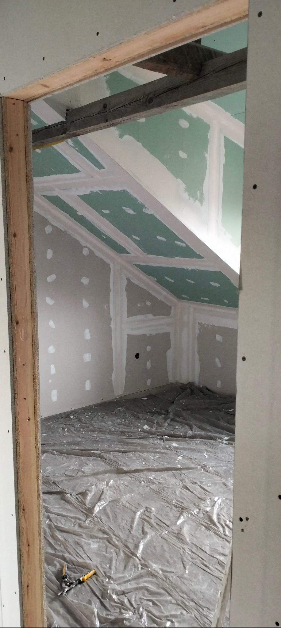 Poddasze w trakcie prac wykończeniowych: płyty gipsowo-kartonowe na skosach sufitu i ścianach, zabezpieczona folią podłoga, widoczne narzędzia. Surowy stan.