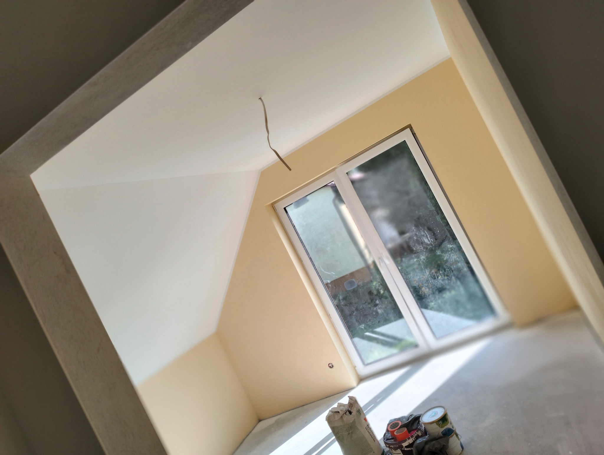 Jasne wnętrze poddasza po malowaniu ścian na beżowo, z białym oknem i widocznymi narzędziami malarskimi na podłodze. Świeżo wykończone ściany i sufit.